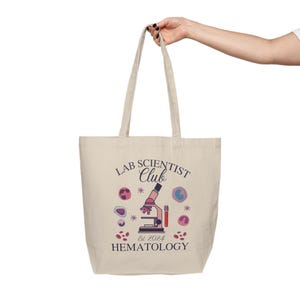 May include: A beige tote bag with a design that says "Lab Scientist Club Est 2024 Hematology". The design also includes a microscope, test tubes, and cells.