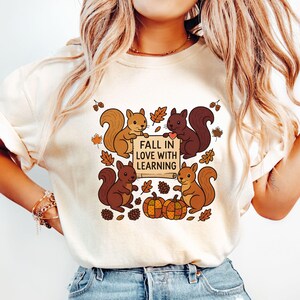 May include: Cream-colored t-shirt featuring a fall-themed graphic. The design includes four cartoon squirrels, autumn leaves, pumpkins, and the text "FALL IN LOVE WITH LEARNING" on a scroll. The squirrels are in shades of brown.