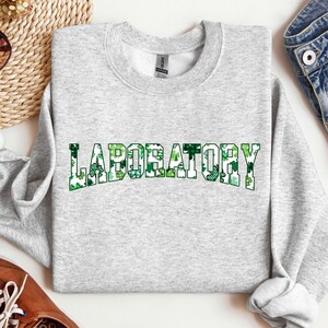 Può includere: Felpa grigio chiaro con la scritta "LABORATORY" in grandi lettere verdi con un motivo a trifogli. La felpa ha un girocollo e maniche lunghe. Il design è a tema San Patrizio.