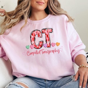Könnte beinhalten: Rosafarbener Pullover mit einem Grafikdruck der Buchstaben "CT" in einem rosa-roten Herzmuster. Die Buchstaben sind von kleinen rosa, roten und orangefarbenen Herzen umgeben. Unter den Buchstaben steht der Text "Computed Tomography".