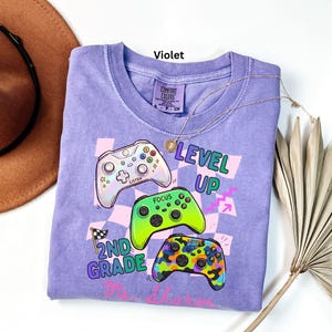 Puede incluir: Una camiseta violeta Comfort Colors con un gr&aacute;fico de tres mandos de videojuegos coloridos y las palabras "LEVEL UP" y "2ND GRADE". Un collar dorado est&aacute; drapeado sobre la camiseta.