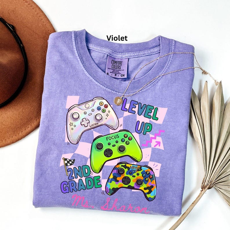 Puede incluir: Una camiseta violeta Comfort Colors con un gr&aacute;fico de tres mandos de videojuegos coloridos y las palabras "LEVEL UP" y "2ND GRADE". Un collar dorado est&aacute; drapeado sobre la camiseta.