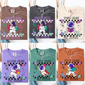 May include: Six different t-shirts with grade level designs. Each shirt features an astronaut graphic with the grade level, such as "Fourth Grade" or "Kindergarten", and "Mrs. Johnson" printed below. The shirts are in various colors.