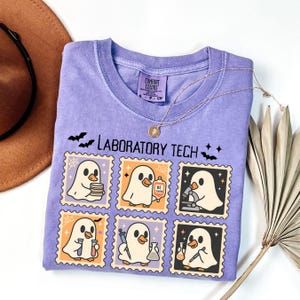 May include: A lavender Comfort Colors t-shirt featuring a "Laboratory Tech" design. The design includes cartoon ghost characters in postage stamp frames, with bats and spiderwebs. A gold necklace is draped over the shirt.