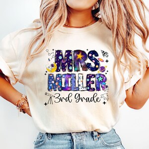 May include: Cream-colored t-shirt with a colorful space-themed design. The design includes the text "MRS MILLER 3rd Grade" in a galaxy-inspired font, along with illustrations of planets, stars, and a rocket ship. A fun and educational teacher shirt.