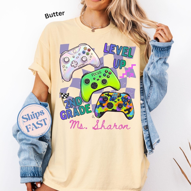 Puede incluir: Camiseta de color mantequilla con tres mandos de videojuegos coloridos. La camiseta tiene las palabras "LEVEL UP", "2ND GRADE" y "Ms. Sharon" impresas. La camiseta tiene un corte holgado.