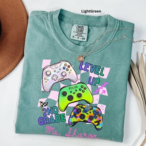 Puede incluir: Camiseta verde claro con un gr&aacute;fico de tres mandos de videojuegos coloridos y las palabras "LEVEL UP", "2ND GRADE" y "Ms. Sharon". La camiseta tiene cuello redondo y mangas cortas. Tambi&eacute;n se ve un collar plateado con un colgante.
