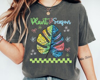 May include: Dark gray t-shirt with a colorful monstera leaf design. The leaf is a mix of yellow, blue, and pink. The text "Plant Season" is above the leaf in green and blue. Small flowers and bows surround the leaf.