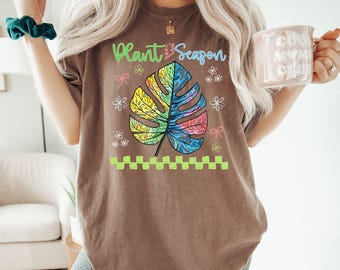 May include: Brown t-shirt with the text "Plant Season" and a colorful leaf design. The leaf is a mix of yellow, green, blue, and pink. The shirt also features small flower and bow graphics.