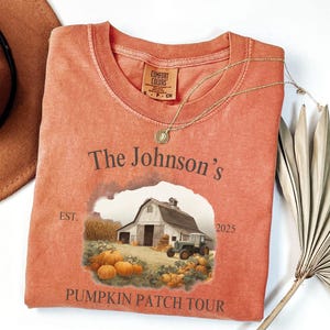 May include: A coral-colored t-shirt with a graphic of a barn, pumpkins, and a tractor. The text reads "The Johnson's Pumpkin Patch Tour" with the year 2025. A gold necklace and a brown hat are also visible.