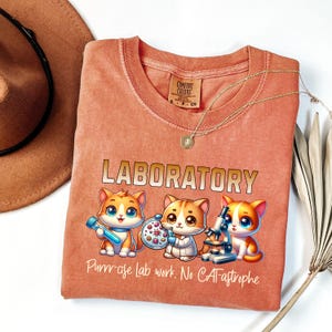 May include: A peach-colored t-shirt with a graphic of three cartoon cats wearing lab coats and holding science equipment. The text "LABORATORY" is above the cats and "Purr-cise Lab work. No CATastrope" is below them.