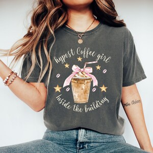 May include: A dark gray t-shirt with a graphic design of a coffee drink with whipped cream and a pink ribbon. The text on the shirt reads "Hypest Coffee Girl Inside the Building".