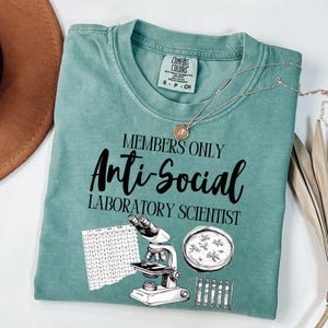 May include: A teal Comfort Colors t-shirt with the text "MEMBERS ONLY Anti-Social LABORATORY SCIENTIST". The shirt features illustrations of a microscope, petri dish, and test tubes. A gold necklace is draped across the shirt.