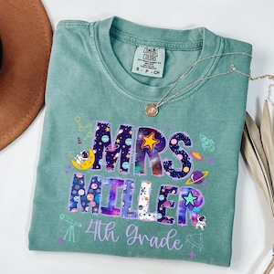 May include: A teal Comfort Colors t-shirt with a space-themed design. The shirt features the text "MRS MILLER 4th Grade" in a colorful, galaxy-inspired font, with illustrations of planets, rockets, and astronauts.