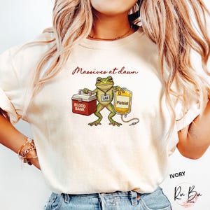May include: Ivory-colored t-shirt featuring a cartoon frog holding a blood bank cooler and a platelet bag. The text "Massives at dawn" is written above the frog. The shirt also has the word "IVORY" and a store logo.