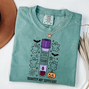 May include: A teal Comfort Colors t-shirt featuring a Halloween-themed graphic. The design includes a purple and white vial with the text "WICKED TOWN Goth Doe", ghosts, bats, a jack-o'-lantern, and the phrase "QUANTITY NOT SUFFICIENT". A gold necklace is draped across the shirt.