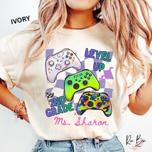 Puede incluir: Sudadera de color marfil con tres mandos de videojuegos coloridos. El dise&ntilde;o incluye las palabras "LEVEL UP", "2ND GRADE" y "Ms. Sharon". El fondo tiene un patr&oacute;n de cuadros. Una prenda casual.