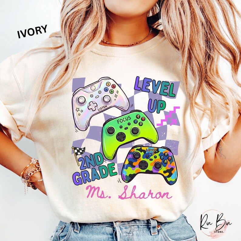 Puede incluir: Sudadera de color marfil con tres mandos de videojuegos coloridos. El dise&ntilde;o incluye las palabras "LEVEL UP", "2ND GRADE" y "Ms. Sharon". El fondo tiene un patr&oacute;n de cuadros. Una prenda casual.