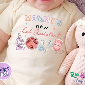 May include: A cream-colored baby bodysuit featuring the text Mommy's new Lab Assistant in colorful lettering. The graphic includes a purple microscope, a petri dish, a blue flask, two pipettes, and a pink blood bag labeled BE POSITIVE.