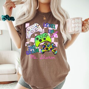 Puede incluir: Camiseta marr&oacute;n con tres coloridos mandos de videojuegos. La camiseta presenta las palabras "LEVEL UP" y "2ND GRADE Ms. Sharon". Los mandos son blancos, verdes y multicolores, sobre un fondo a cuadros.