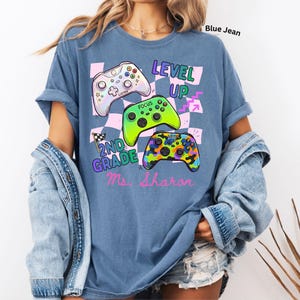 Puede incluir: Camiseta azul vaquero con un dise&ntilde;o gr&aacute;fico que presenta tres mandos de videojuegos coloridos y las palabras "LEVEL UP" y "2ND GRADE Ms. Sharon". El dise&ntilde;o incluye un fondo a cuadros.