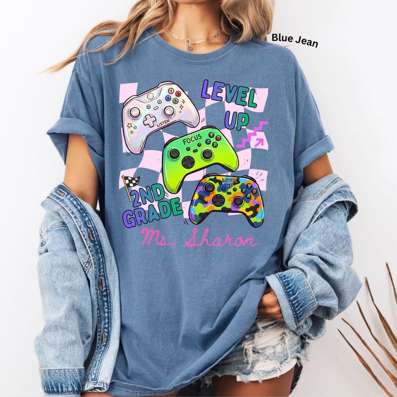 Puede incluir: Camiseta azul vaquero con un dise&ntilde;o gr&aacute;fico que presenta tres mandos de videojuegos coloridos y las palabras "LEVEL UP" y "2ND GRADE Ms. Sharon". El dise&ntilde;o incluye un fondo a cuadros.