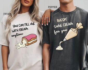 May include: Ivory and pepper-colored t-shirts with humorous graphics. The ivory shirt features a cartoon pastry with the text "You can fill me with cream anytime." The pepper shirt has a pastry bag graphic and the text "Bussin' some cream for you."
