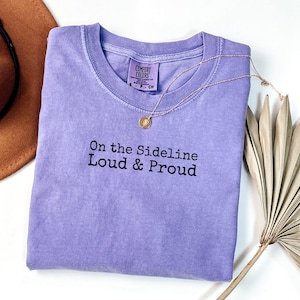 May include: A lavender colored t-shirt with the text "On the Sideline Loud & Proud" printed in black.