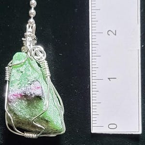 May include: A pendant featuring a rough-cut green stone with red and black inclusions, wrapped in silver-colored wire. The stone is approximately 1 inch in length. The pendant hangs from a delicate chain.