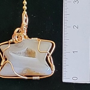 Könnte beinhalten: Ein golddrahtumwickelter Anhänger mit einem weißen Achat-Stein mit braunen Einschlüssen. Der Anhänger ist etwa 2,5 cm groß.