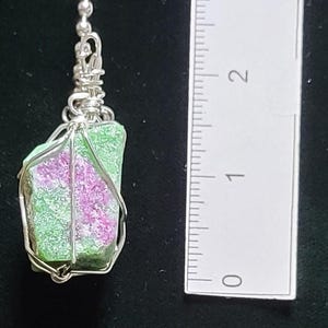May include: A pendant featuring a rough-cut gemstone with green and pink hues, encased in a silver wire wrap. The pendant is suspended from a delicate silver chain. A ruler is placed next to the pendant for scale.