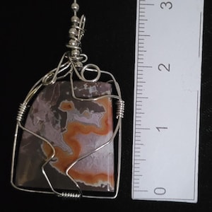 Puede incluir: Colgante artesanal con una piedra rectangular y alambre de plata. La piedra muestra tonos grises, naranjas y blancos. El colgante mide aproximadamente 2,5 cm de largo. Una joya única.