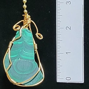 May include: A malachite pendant, approximately 1.5 inches long, is wire-wrapped in gold-colored metal. The pendant hangs from a gold-colored chain. The malachite stone displays a swirling pattern of dark and light green hues.