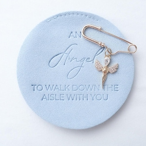 Pode incluir: Um alfinete de segurança dourado com um pingente de anjo com uma pedra preciosa transparente e pequenos detalhes brilhantes. O alfinete está sobre uma superfície circular azul claro com o texto em relevo "AN Angel TO WALK DOWN THE AISLE WITH YOU."
