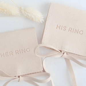 May include: Two square, beige fabric pouches with ribbon ties at the bottom. One pouch is embossed with the text HER RING, and the other pouch is embossed with the text HIS RING. The pouches have a soft, suede-like texture and a neutral color.