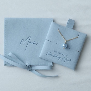 Op de afbeelding: Een lichtblauwe sieraden cadeau set. Het bevat een gouden ketting met een blauwe parel hanger. De set bevat een zakje met het woord "Mom" in reliëf en een ander zakje met de tekst "Will you be my Something Blue?"