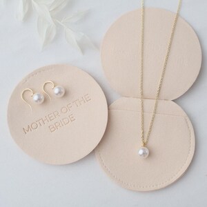 May include: A gold necklace with a single pearl pendant and a pair of gold pearl earrings. The earrings and necklace are displayed on a light pink circular holder with the words "MOTHER OF THE BRIDE" embossed on it.
