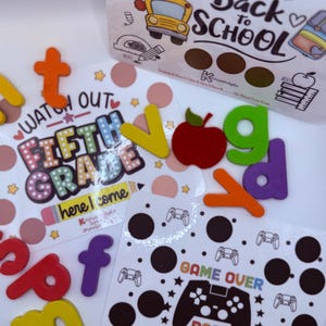May include: A collection of colorful school-themed items, including a box with "Back to School" text and a school bus illustration. There are also cards with "Fifth Grade" and "Game Over Back to School" text, along with colorful letter cutouts and an apple.