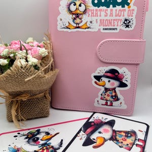 May include: A pink planner with a snap closure, adorned with duck-themed stickers. The stickers feature cartoon ducks in hats and coats, and text that reads "QUACK! THAT'S A LOT OF MONEY!" A small bouquet of pink and white flowers is in the background.