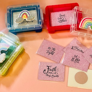 May include: Assortment of small clear plastic boxes with colorful rainbow and cloud designs and inspirational quotes on pink glitter paper. The quotes include "Love Each Other", "Be The Light", "Faith Makes All Things Possible", "Let It Be", "Be Thankful", and "Choose Joy".