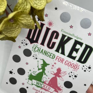 Puede incluir: Una tarjeta para rascar blanca con la palabra "WICKED" en negro y rosa, con ilustraciones de una bruja y un hada. La tarjeta tiene círculos y estrellas negros, con el texto "CHANGED FOR GOOD".