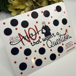 May include: A white rectangular sticker with black polka dots and red accents. The text "NO! that wasn't a Question" is written in red and black script. The sticker features an illustration of a woman in sunglasses and the words "THE DEVIL WEARS PRADA".