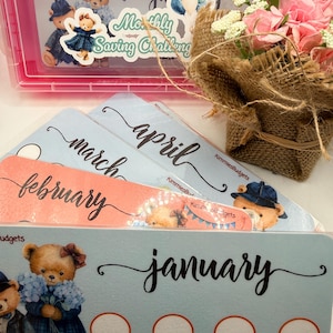 May include: A pink plastic box with a "Monthly Saving Challenge" sticker and several budget cards. The cards are labeled with months and dollar amounts. Decorative teddy bear stickers are also present.