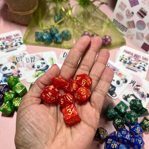 Op de afbeelding: Een hand die een set rode dobbelstenen met gouden nummers vasthoudt. De dobbelstenen liggen op een roze oppervlak met andere dobbelstenen in blauw, groen en zwart.