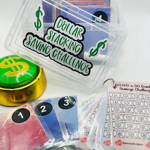 Może przedstawiać: Przezroczyste plastikowe pudełko z zielonym znakiem dolara i tekstem "Dollar Stacking Saving Challenge" na pokrywie. Pudełko zawiera stos kopert z numerami. Są też trzy koperty z numerami 1, 2 i 3. Koperty leżą na białym tle.