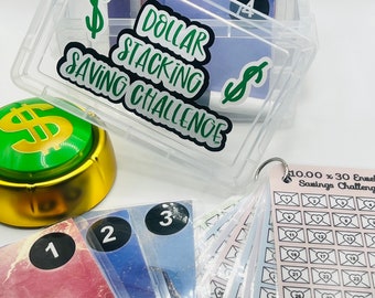 May include: A clear plastic box with a green dollar sign and the text "Dollar Stacking Saving Challenge" on the lid. The box contains a stack of envelopes with numbers on them. There are also three envelopes with numbers 1, 2, and 3 on them. The envelopes are on a white surface.