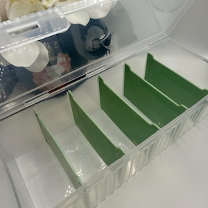 May include: A clear plastic storage container with a hinged lid and several green dividers. The container is rectangular and has a ridged design along the sides. The dividers create separate compartments.