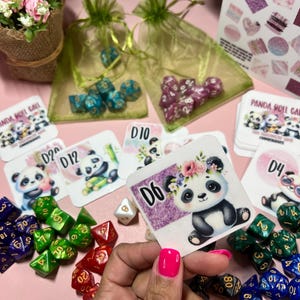 Op de afbeelding: Een set kleurrijke dobbelstenen met een witte dobbelsteen en een kaart met een panda-illustratie. De kaart heeft de tekst "D6" erop. De kaart heeft een paarse achtergrond met witte bloemen en een roze rand. De dobbelstenen liggen op een roze oppervlak.