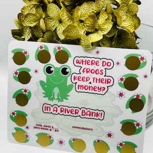 May include: A white savings tracker card with a green frog illustration and the text "Where do frogs keep their money? In a river bank!" The card has gold scratch-off circles and small pink and white flower accents.