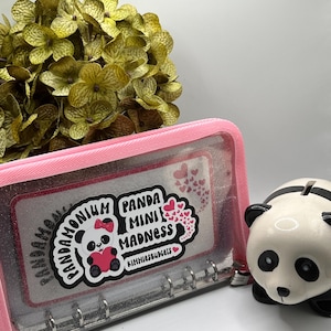 May include: A pink zippered binder with a clear front panel featuring a sticker that says "Panda Mini Madness" with a panda bear graphic. A white and black panda piggy bank is to the right.